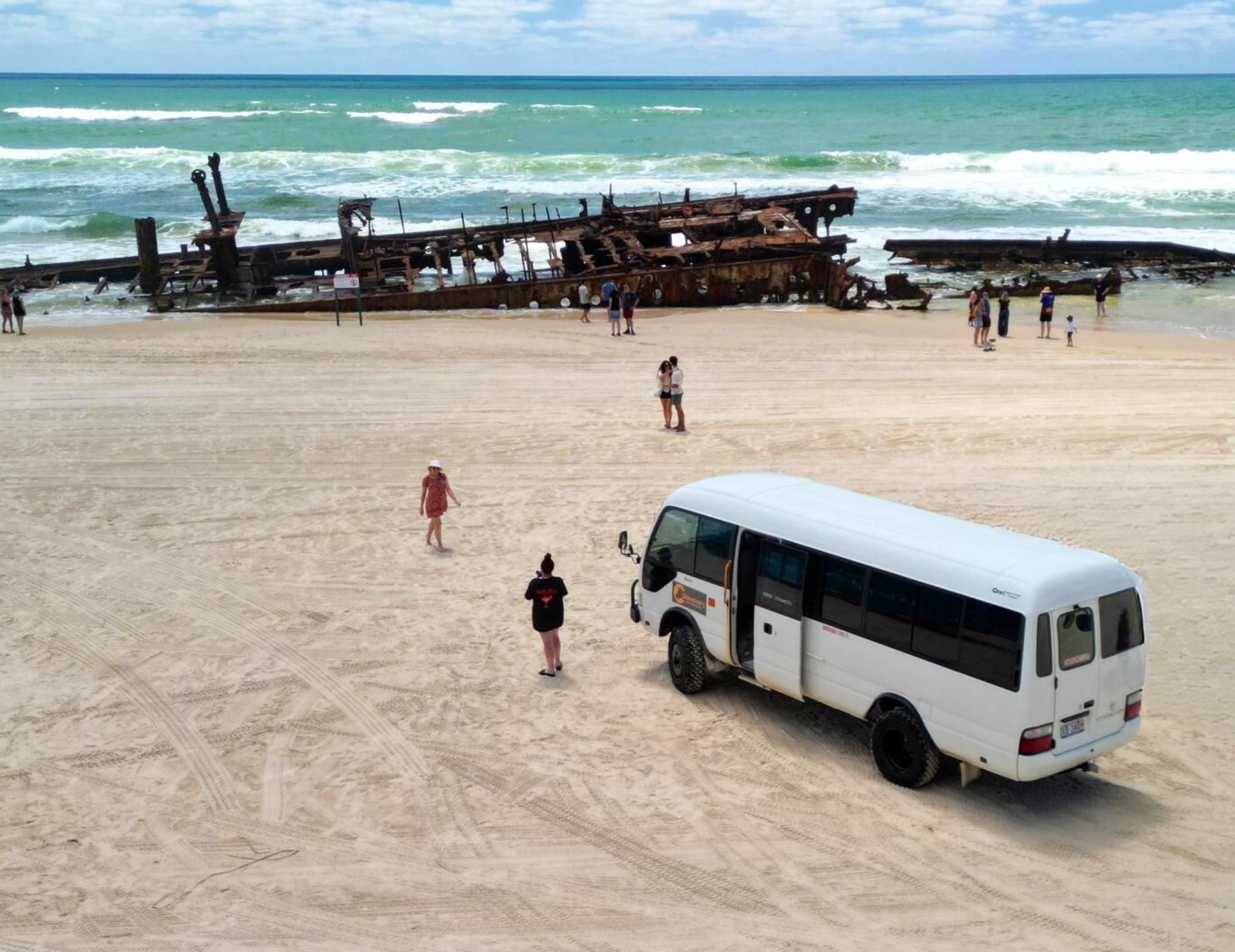 White coach parked on sandy Fraser Island beach by holidaymakers and iconic rusted shipwreck, K'gari Day Tour Ex Noosa, Australia.