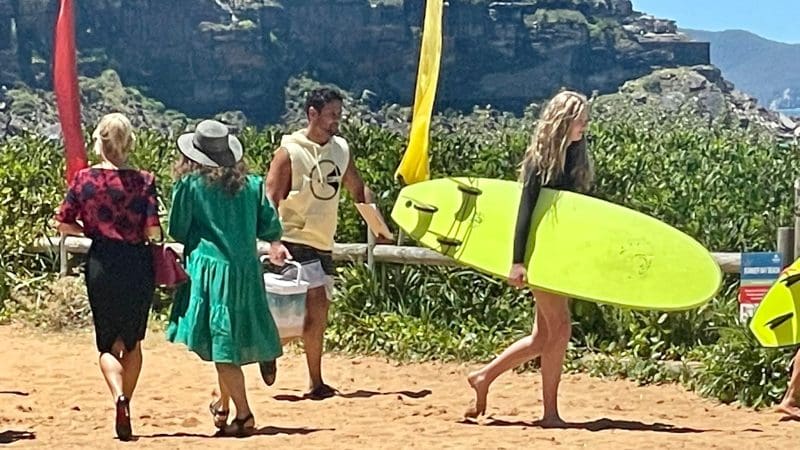 Four people walk along a sandy path, filming possibly for Celebtime Tours or 1 Day Home Away Tour, with a bright yellow surfboard nearby.