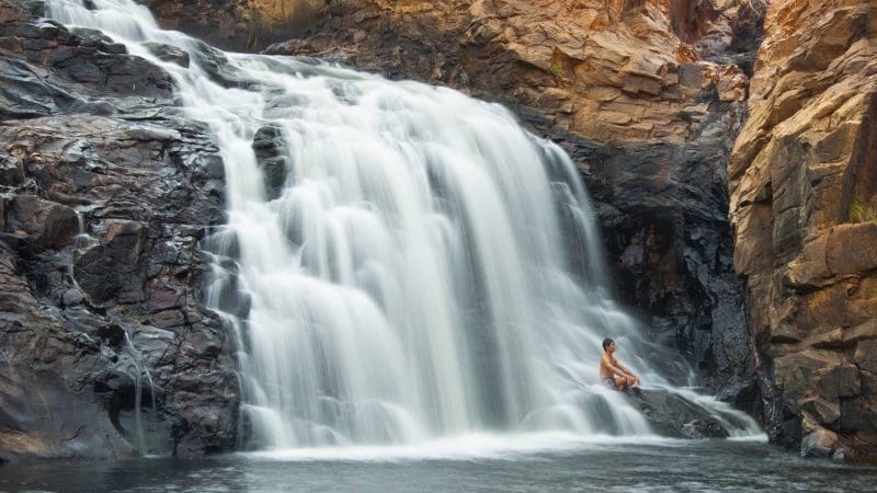 Traveller relaxing on sunlit rocks beside a stunning cascading waterfall during the 10 Day Broome to Darwin 4WD Kimberley Adventure.