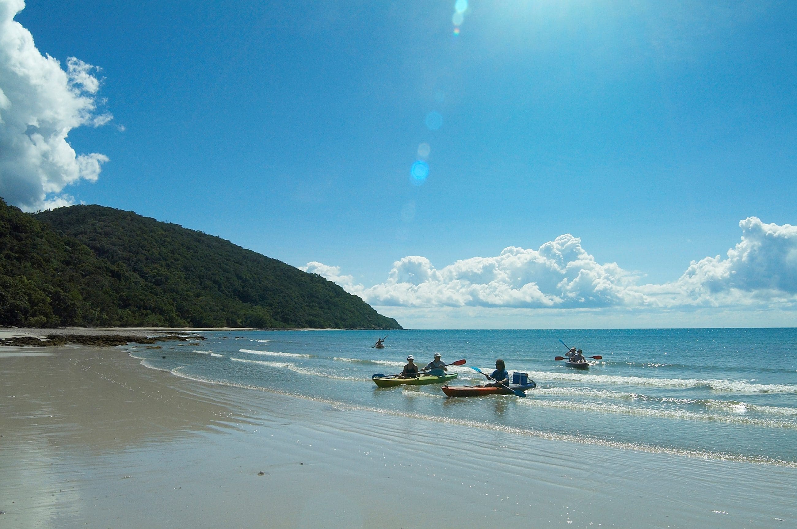 Adventurers kayaking by the Cape Tribulation coast under clear skies, lush green hills in the background—top-rated tropical tour scene.