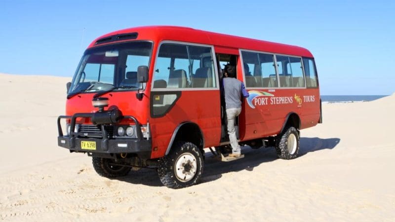 Vibrant Collective Travel coach for 1 Day Whale Watching Port Stephens parked on sandy ground as passenger boards, ready for adventure.