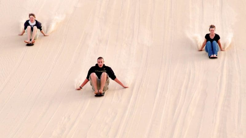 Three adventurers sandboard side by side down Port Stephens dunes with Colourful Collective Travel, experiencing an epic racing adventure.