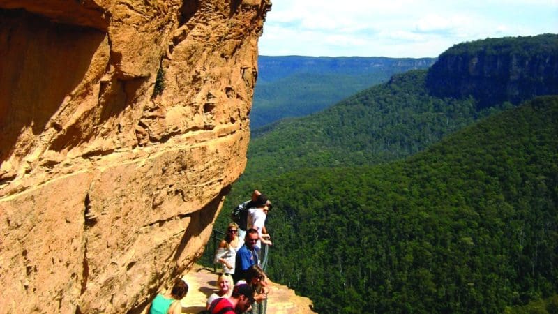 Adventurous hikers traverse a narrow cliffside trail on a 1 Day Blue Mountains Wildlife Park Tour, with breathtaking valley views.
