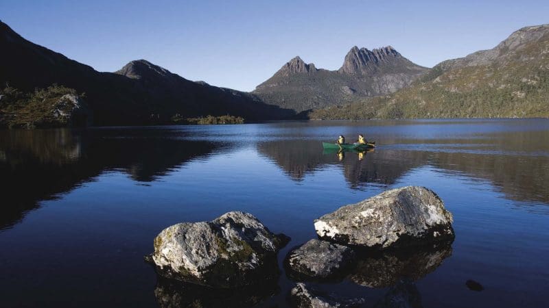 Adventurers canoe on a serene Tasmanian lake surrounded by large rocks, showcasing the 6 Day Tasmania Explorer Tour experience.