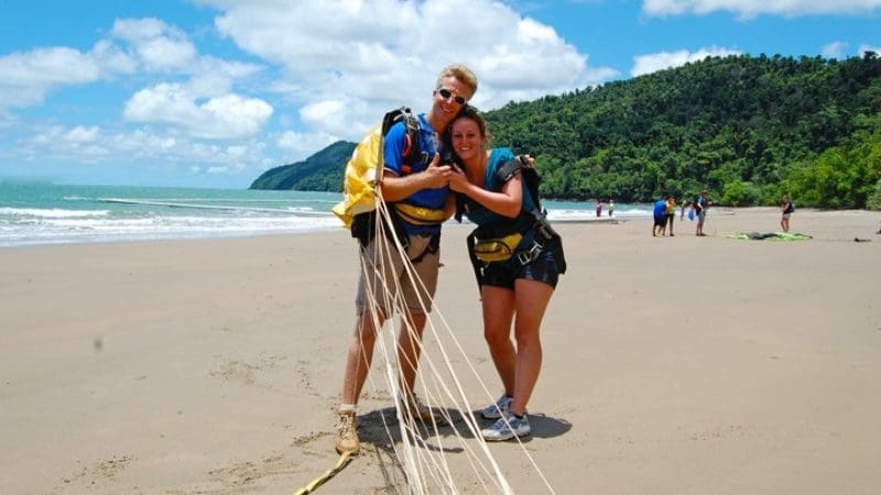 Two harnessed skydivers smile on Sydney’s sandy beach, celebrating their Tandem Skydive Extras beneath a bright blue sky.