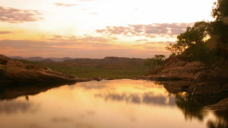 Serene lake mirrors soft pastel sunset sky, highlighting Kakadu Tour Highlights with rugged rocks and sparse shoreline trees.