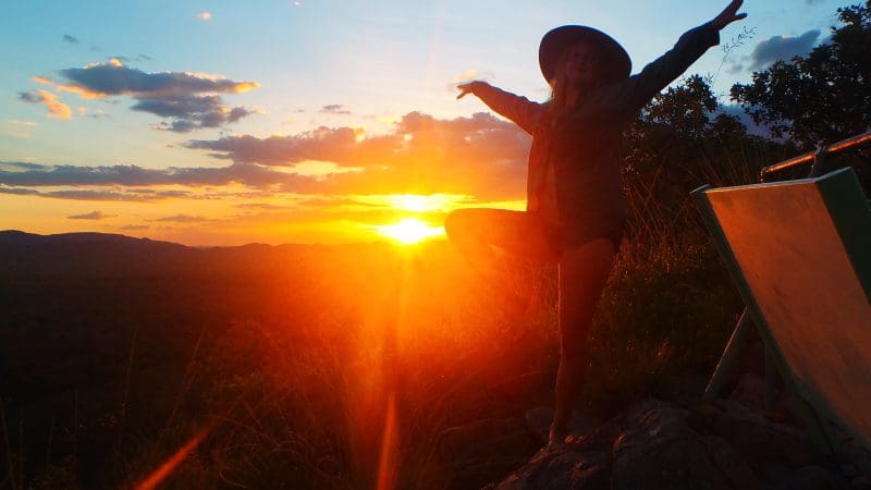 Traveller in a wide-brimmed hat strikes a yoga tree pose atop a hill at sunset, showcasing Kakadu Tour highlights and vibrant skies.