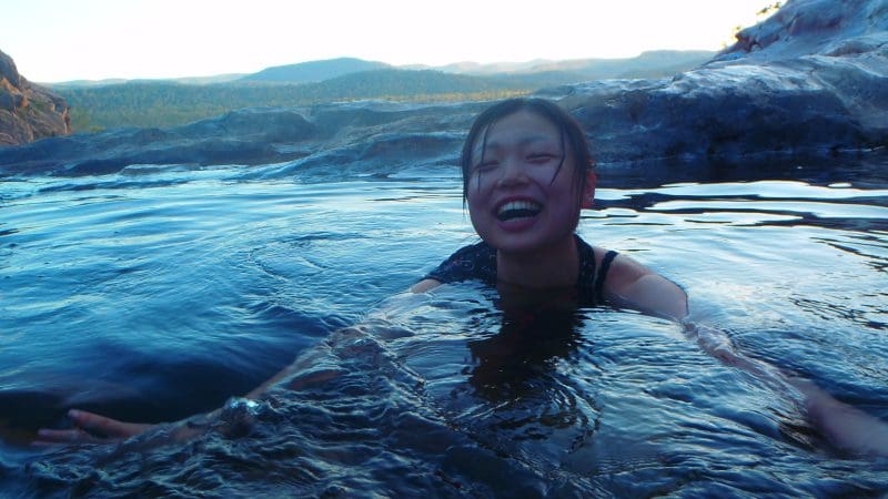 Smiling woman swimming in crystal-clear Kakadu waters at sunset, framed by hills and rock formations—a top-rated tour highlight.