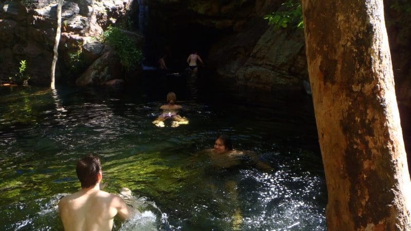 Four people swim in a picturesque, shaded rocky pool beside a cave, surrounded by lush trees—top Kakadu National Park attraction.