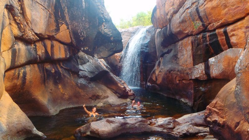 Visitors swim and unwind in a scenic natural pool at Kakadu, framed by striking cliffs and a picturesque cascading waterfall.