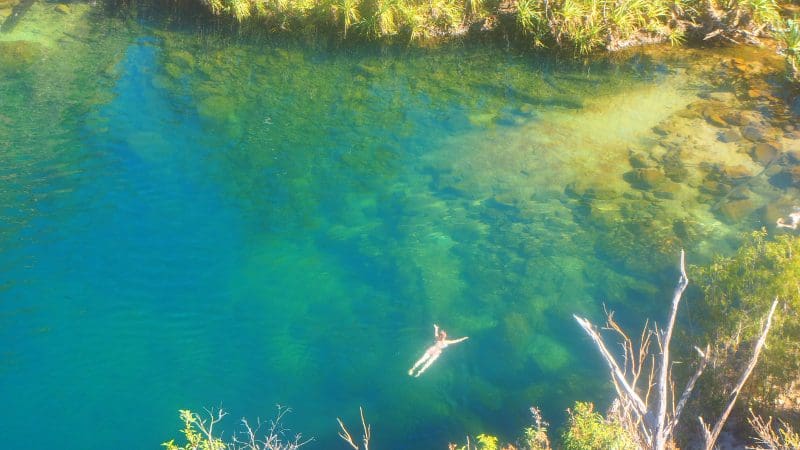 Experience a Kakadu Tour as a person floats in crystal-clear blue river waters by rocky banks, immersed in lush green Australian scenery.