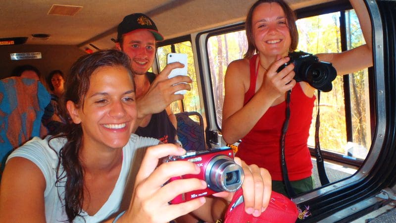 Three tourists smiling on a Kakadu National Park tour capture photos from their safari vehicle, enjoying a sunny day before heading to their hotel.