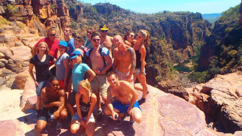 Tour group stands atop rocky cliffs on a Kakadu adventure, overlooking scenic canyon, winding river, lush trees, and bright blue sky.