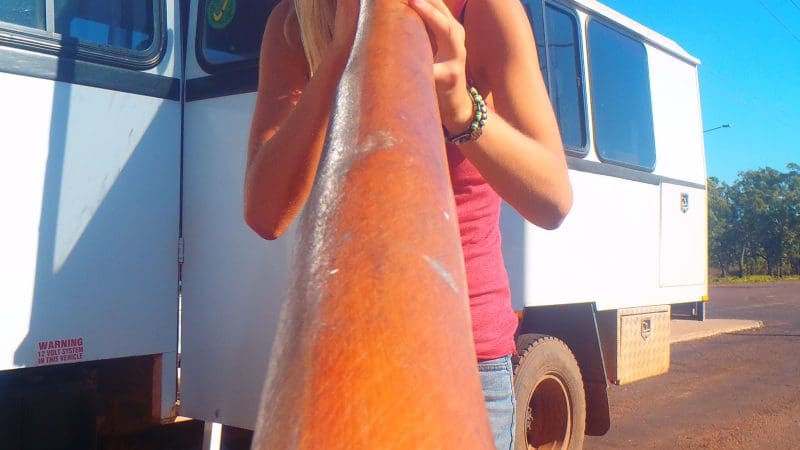 Woman in sunglasses plays didgeridoo near a white 4WD on a sunny Kakadu Adventure Tour, showcasing Outback culture and travel experience.
