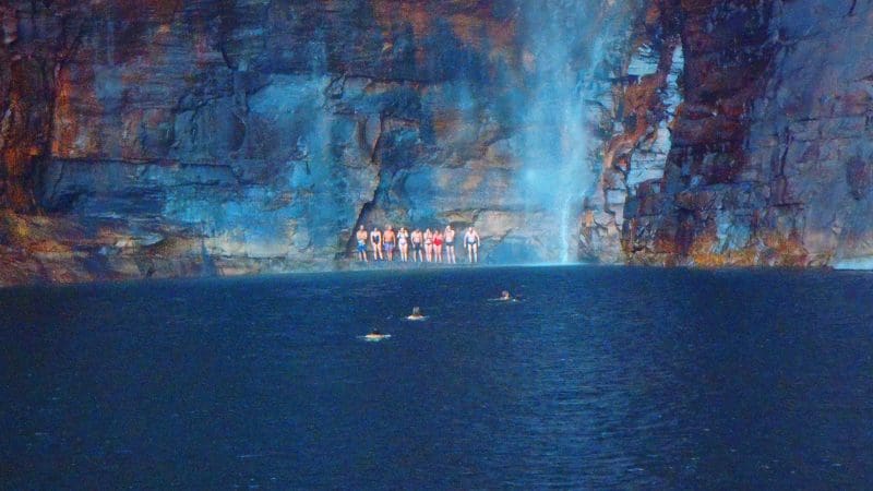 Tourists on a Kakadu guided tour gather under a stunning waterfall, as visitors swim in crystal-clear blue water near a nearby hotel.