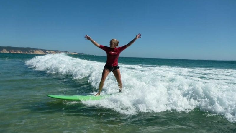Surfer in a red shirt rides a gentle wave at Learn To Surf Australia’s Longest Wave, arms wide open under clear blue skies.