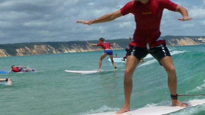 Surfers riding crystal-clear turquoise waves near scenic cliffs, with a Double Island Surfboard Hire 4x4 Adventure vehicle and group nearby.