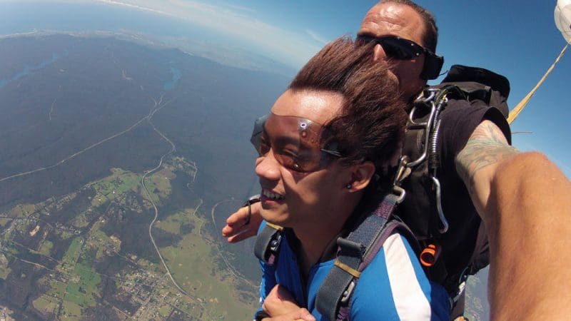 Experience an exhilarating Skydive Sydney tandem jump, soaring above lush green fields and forests under a stunning clear blue sky.