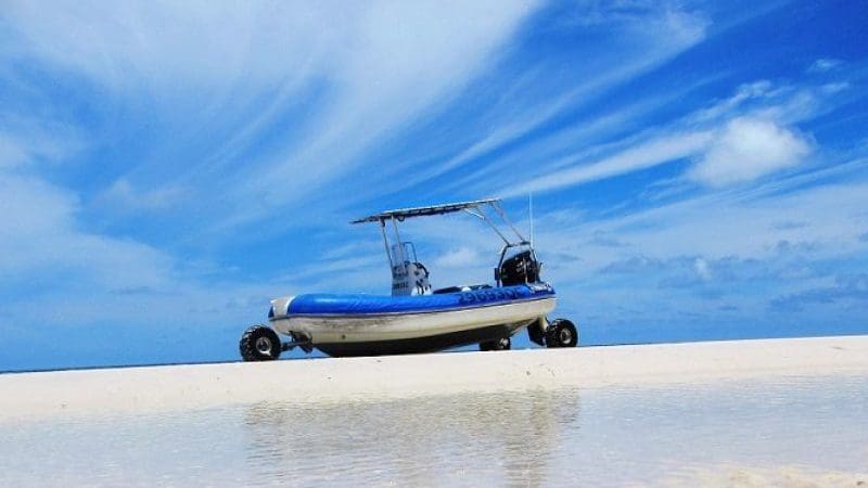 A blue inflatable boat on pristine white sand awaits a Remote 1 Day K'gari Fraser Island tour beneath a vivid, inviting sky.