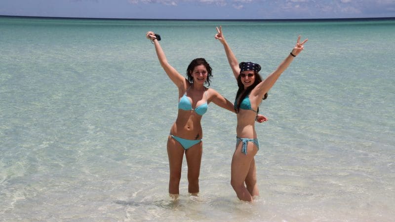 Two women in matching blue bikinis joyfully celebrate in crystal-clear sea water on a Remote 1 Day K'gari Fraser Island tour experience.