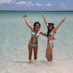 Two women in blue bikinis raise their arms whilst enjoying a Remote 1 Day Kgari Fraser Island tour in crystal-clear sea waters.