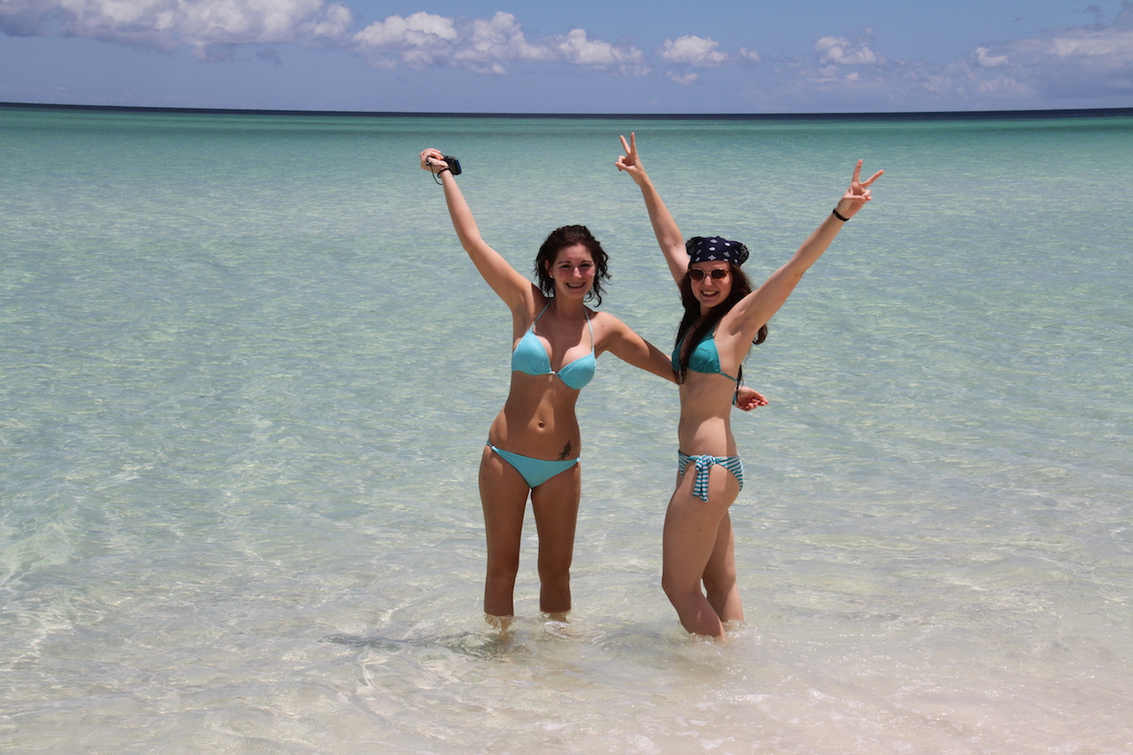 Two women in blue bikinis raise their arms whilst enjoying a Remote 1 Day Kgari Fraser Island tour in crystal-clear sea waters.