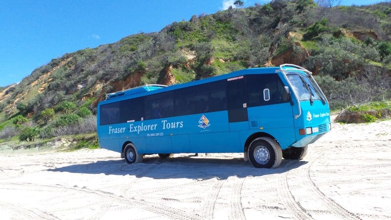 Fraser Explorer Tours blue bus on sandy K'gari beach, featured in the 4 Day Experience, with lush green hills in the background.