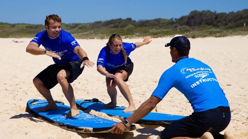 Two people master surf stances on Byron Bay’s sunny beach during a Learn to Surf Half-Day Tour, perfect for beginners and adventure seekers.
