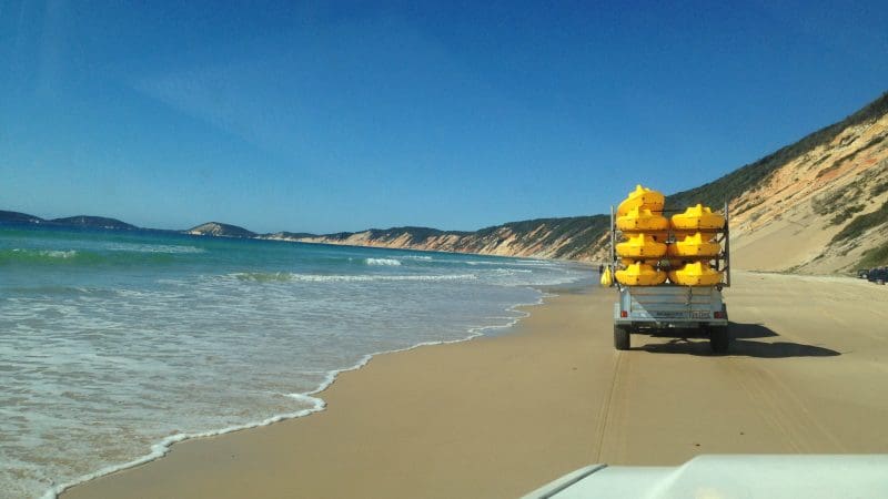 Yellow Dolphin Kayaking gear on a trailer towed down Rainbow Beach, bordered by turquoise ocean waves and dramatic rocky cliffs.