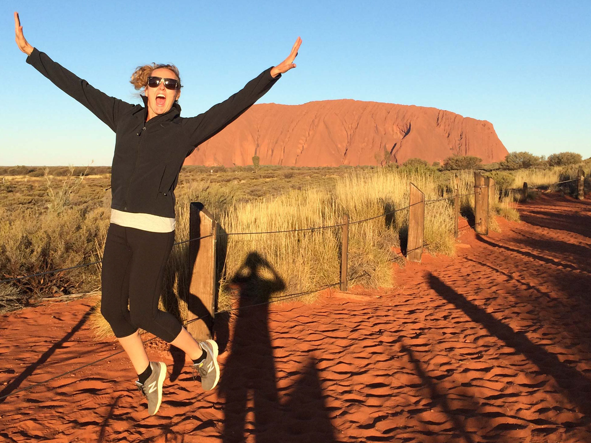 Excited woman leaps with arms raised on red dirt track in Macdonnell Ranges near Uluru, under vibrant blue sky, Australia adventure.