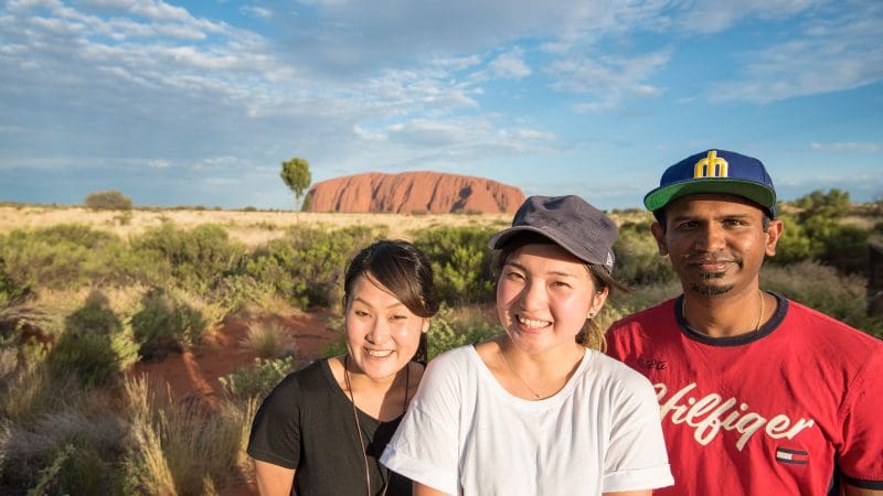Three happy travellers smile together at Uluru, enjoying the iconic 3 Night Rock The Centre Yulara to Yulara adventure tour outdoors.