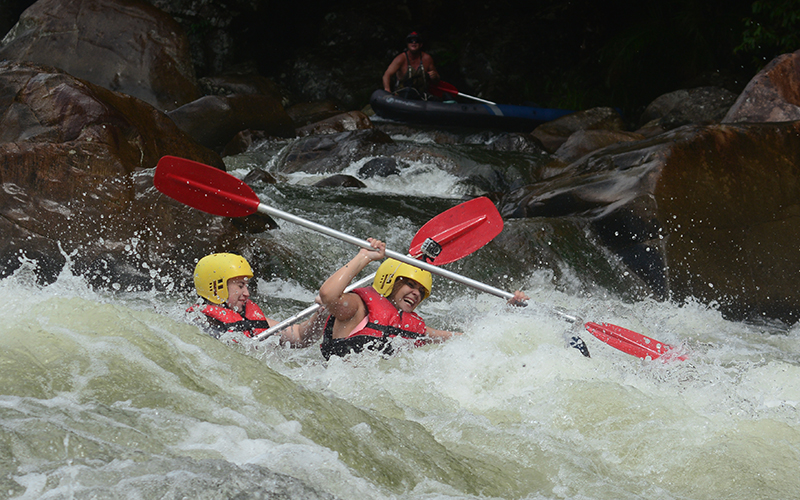 Adventurous duo in helmets kayaking fierce rapids during a Half Day Sports Rafting trip, paddles lifted in thrilling excitement.