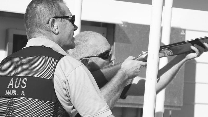 Two men at Vic Werribee shooting range, one coaching while the other aims a shotgun. Vest says 