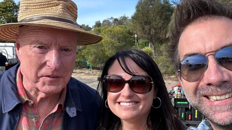 Three happy travellers enjoy a 1 Day Home Away Tour outdoors, wearing sunglasses and a straw hat, with trees and a clear blue sky.