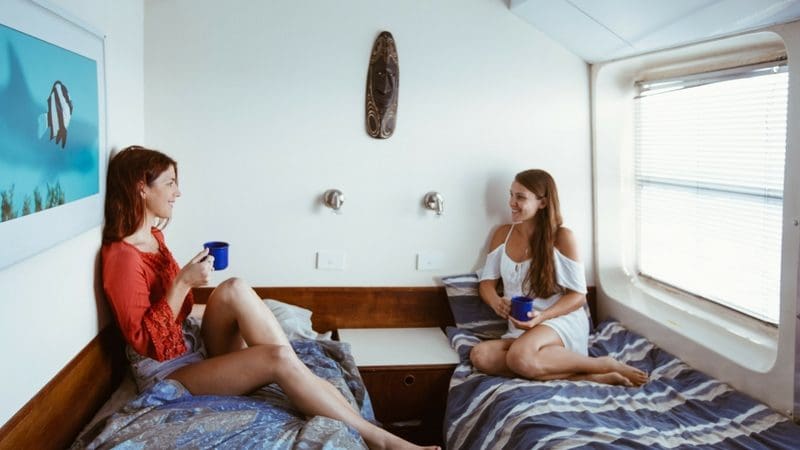 Two women relax on separate beds, smiling and chatting over blue mugs after completing a 6-Day PADI Open Water Advanced Course.