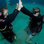 Two scuba divers celebrate with an underwater high five during a 1–2 Day PADI Refresher Course as coursemates watch from the sea bed.