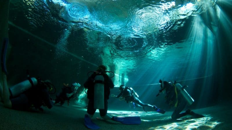 Scuba divers kneel underwater in a clear pool, participating in a 1–2 day PADI refresher course with sunlight streaming down.