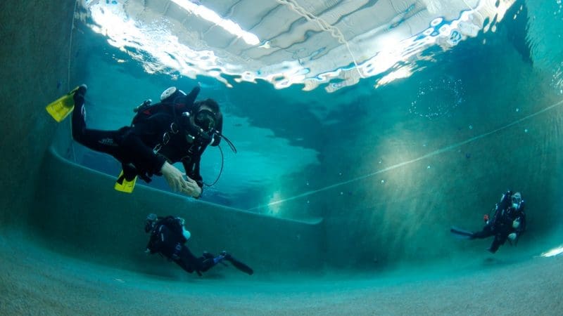 Three scuba divers train underwater in a crystal-clear pool, visible pool walls and ceiling above, during a 1 Day PADI Refresher Course.