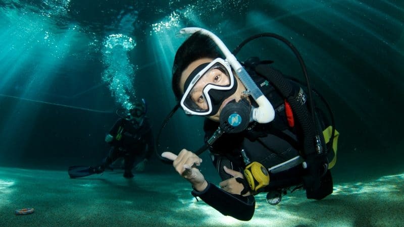 Two scuba divers beneath clear water; one signals thumbs-up during a PADI Refresher Course as sunlight beams illuminate the scene.