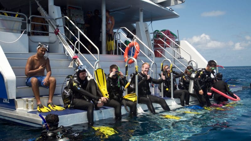 Scuba divers in PADI Open Water Course 2 sit on a boat’s edge, fully equipped with fins and kit, preparing to dive into the sea.