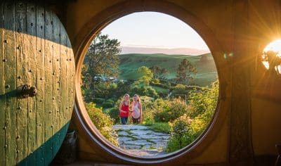 Scenic view through a round door to two people outside in a vibrant, sunlit landscape with green hills, lush gardens, and blue sky.