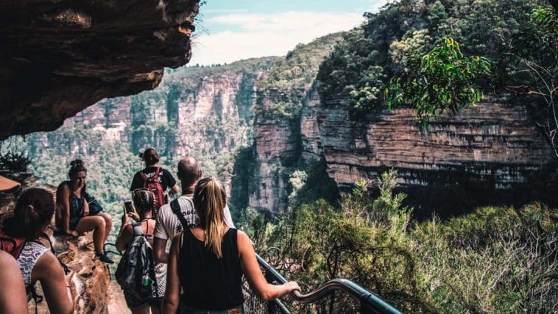 Walkers on a Blue Mountains day tour walk a scenic rocky path above a gorge, surrounded by lush views, sharing a picnic lunch.