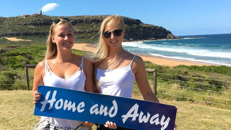 Two women in white tops display a Home and Away sign on Celebtime Tours beach, with cliffs and blue sky in the sunshine behind them.