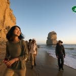 Four travellers stroll along a sandy beach at sunset by scenic cliffs on a Great Ocean Road 1 Day Sunset Tour; Bunyip Tours logo visible.