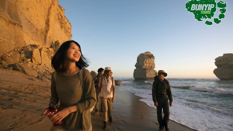 Four travellers stroll along a sandy beach at sunset by scenic cliffs on a Great Ocean Road 1 Day Sunset Tour; Bunyip Tours logo visible.