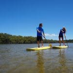 Experience a 5-hour Byron Bay stand up paddle board tour with two people gliding along a tranquil river beneath sunny blue skies.
