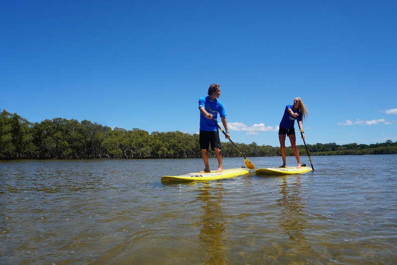 Experience a 5-hour Byron Bay stand up paddle board tour with two people gliding along a tranquil river beneath sunny blue skies.