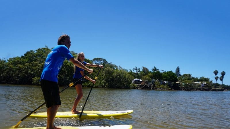 Two people paddleboard on calm waters during the Byron Bay Group 2 Five Hour Stand Up Paddle Board Tour under clear sunny skies.