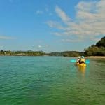 Kayaking duo paddles the scenic Brunswick River Nature Tour, framed by vibrant green trees and a stunning, clear blue sky above.