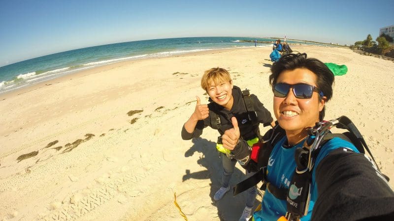 Two smiling skydivers in full kit give thumbs up after a Sunshine Coast Beach tandem skydive with blue skies and sea views.